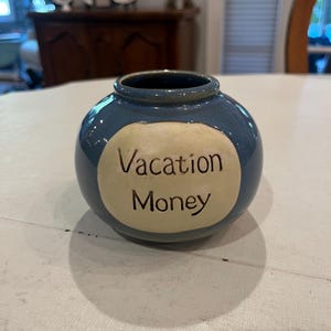 May include: A small, round, blue ceramic jar with a cream-colored oval patch. The words "Vacation Money" are written in black script on the patch. The jar has a dark blue interior and sits on a white surface.