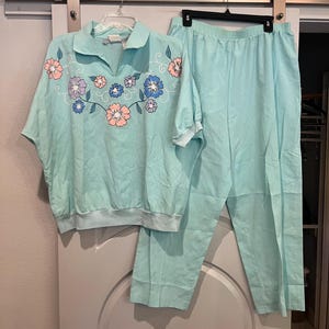 May include: A light blue two-piece leisure suit. The top has a collared neckline and short sleeves, adorned with embroidered flowers in pink, purple, and blue. The pants are a matching light blue color with an elastic waistband. The suit is hanging on a hanger.