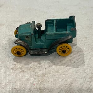 May include: A vintage teal toy car with yellow wheels. The toy car has a detailed design, including a steering wheel and a rear cargo area. The wheels are a bright yellow colour, contrasting with the teal body. The car appears to be made of metal.