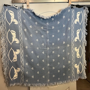 May include: A light blue woven throw blanket featuring a repeating pattern of white rocking horses along the vertical side borders and a central field filled with small white hearts. The blanket has a textured fringe along all four edges.