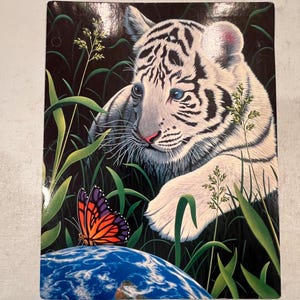 Puede incluir: Una impresión rectangular que presenta un cachorro de tigre blanco con rayas negras, ojos azules y nariz rosa, sobre un fondo de hierba verde y un fondo oscuro. Una mariposa colorida descansa sobre un globo azul. Tema de fantasía.