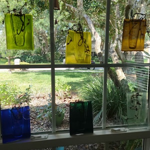 May include: Six glass vases of different colors, hanging in a window. The vases are clear, green, blue, and yellow. They are all hanging from hooks on the window frame. The vases are empty.
