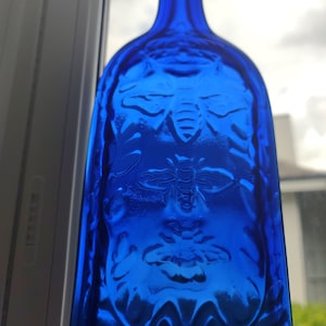Op de afbeelding: Een kobaltblauwe glazen fles met een reliëf van bijen. De fles is ondoorzichtig en heeft een ronde vorm.