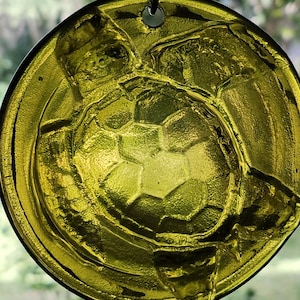 May include: A round, green glass ornament with a raised design of a sea turtle. The ornament has a hole at the top for hanging.