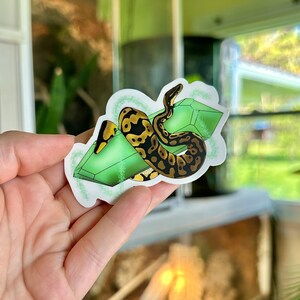 Op de afbeelding: Een sticker met een groene en zwarte slang die om een ​​groen kristal is gewikkeld. De slang heeft een geel en zwart patroon. De sticker is op een witte achtergrond.