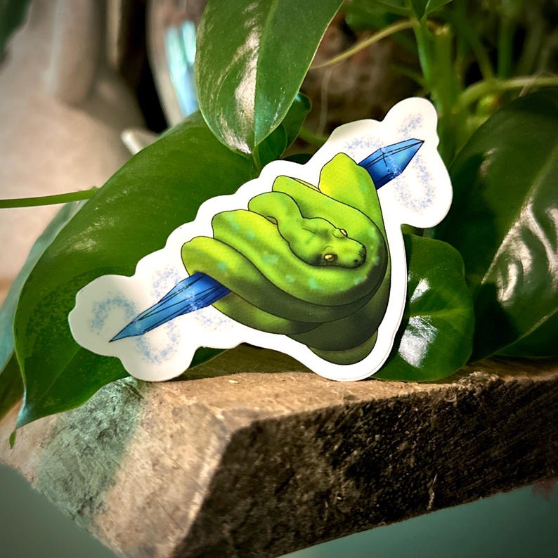 May include: A sticker featuring a green snake coiled around a blue crystal. The snake has a detailed, textured appearance with darker green shading. The sticker has a white border with light blue accents, set against a backdrop of green leaves and a wooden surface.
