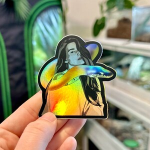 Op de afbeelding: Een holografische sticker met een vrouw met lang donker haar en een slang die om haar heen is gewikkeld. De sticker heeft een regenboogkleurige, iriserende afwerking.