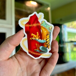 May include: A sticker featuring a green and yellow frog clinging to a red crystal. The crystal is surrounded by a golden halo.