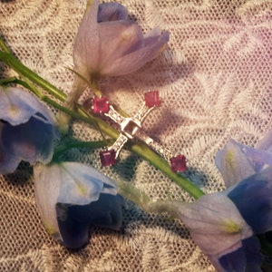 Puede incluir: Un colgante de cruz de plata con cuatro piedras preciosas rosas engastadas en el centro de cada brazo. La cruz está sobre un tejido de encaje blanco con flores azules.