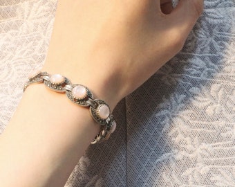 Puede incluir: Un brazalete de plata con una cadena delicada y piedras rosas de forma ovalada. Las piedras están engastadas en plata y tienen un acabado ligeramente iridiscente.