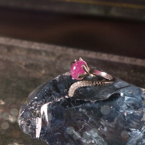 May include: A silver ring with a pink gemstone set in a floral design. The ring is set on a blue and gray rock.
