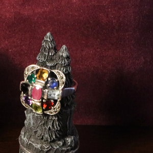 May include: A silver ring with a cluster of colorful gemstones set in a decorative design. The ring is displayed on a small, gray, tree-shaped stand.