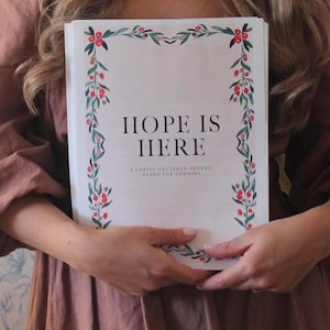 May include: A book titled "HOPE IS HERE" is held by a person. The cover features a decorative border of green leaves and red berries. The text "A CHRIST-CENTERED ADVENT STUDY FOR FAMILIES" is below the title. The book is white with black text.