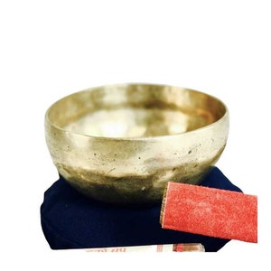 May include: A golden singing bowl with a textured surface, resting on a navy blue cushion. A red suede-like striker and a white and red printed stick are included. The bowl is a traditional meditation tool.