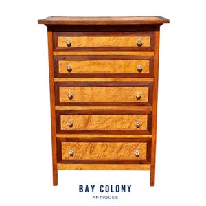 May include: A wooden chest of drawers with five drawers. The drawers have crystal knobs and are framed by darker wood. The chest has a light wood grain pattern on the drawer fronts. The text "BAY COLONY ANTIQUES" is at the bottom.