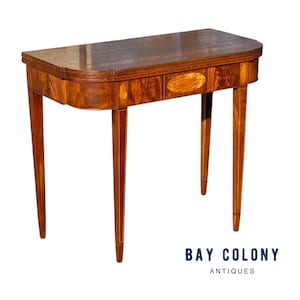May include: A rectangular antique table with a curved front edge. The table is made of dark wood with inlaid details and slender, tapered legs. The table has an oval inlay on the front. The table is a classic piece of furniture.
