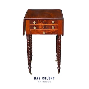 May include: A mahogany antique side table with two drawers and brass knobs. The table has a drop-leaf design and stands on intricately carved, spiral-turned legs. The table is from Bay Colony Antiques.