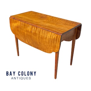 May include: A wooden drop leaf table with a natural wood finish. The table has a rectangular top with rounded edges and four legs. The table is in a folded position, with the leaves down.