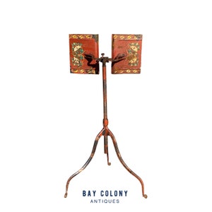 May include: An antique music stand with a red painted finish and intricate floral designs. The stand has a tripod base and two adjustable music rests.