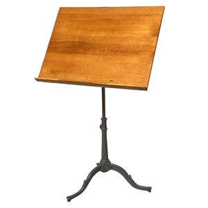 May include: A vintage wooden music stand with a light brown, angled tabletop. The stand has a dark gray, cast iron base with three curved legs. The height-adjustable stand is designed to hold sheet music or books.