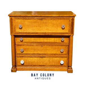 Puede incluir: Un antiguo cofre de cajones de madera con cuatro cajones y un cajón superior con una tapa abatible. El cofre tiene un acabado en madera clara con un patrón de incrustación decorativo. Los cajones tienen tiradores decorativos con pomos de porcelana blanca. El cofre está etiquetado como "Bay Colony Antiques".