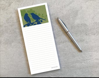 Puede incluir: Un bloc de notas rectangular con líneas horizontales y un gráfico superior que muestra dos pájaros azul oscuro posados en una rama sobre un fondo verde con un sutil patrón de puntos, acompañado de un bolígrafo retráctil plateado.