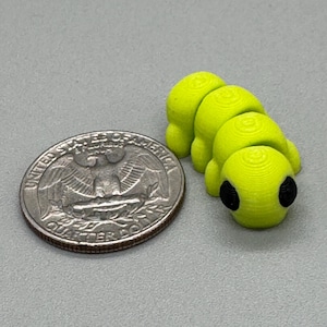 Puede incluir: Un pequeño juguete de oruga impreso en 3D, de color amarillo brillante, con ojos negros. La oruga está junto a una moneda de cuarto de dólar estadounidense para comparar el tamaño. El juguete está hecho de un material texturizado.