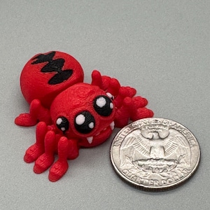 Puede incluir: Figura de araña roja impresa en 3D con ojos grandes y de dibujos animados y una moneda de veinticinco centavos para comparar el tamaño. La araña tiene un cuerpo y patas texturizados, y un diseño negro en la espalda.