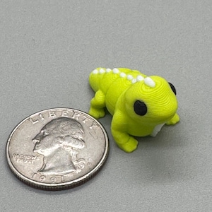 May include: A small, lime green lizard figurine with black eyes and white spots along its back. The figurine is next to a U.S. quarter coin for size comparison. The lizard is on a gray surface.
