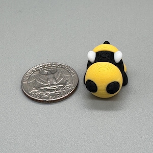Puede incluir: Una pequeña figura de abeja impresa en 3D, amarilla y negra, con antenas blancas. La abeja está junto a una moneda de veinticinco centavos estadounidense para comparar el tamaño. La figura tiene un cuerpo redondeado y descansa sobre una superficie gris.