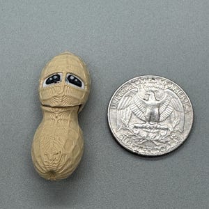 Puede incluir: Una figura de cacahuete beige con una cara triste, junto a una moneda de veinticinco centavos estadounidense. El cacahuete tiene una superficie texturizada y ojos negros. La moneda mide aproximadamente 2,4 cm de diámetro.