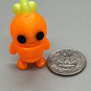 May include: A small, orange figurine resembling a cartoon character with black eyes and a green crown. The figurine is next to a U.S. quarter for size comparison. The character has small arms and legs.