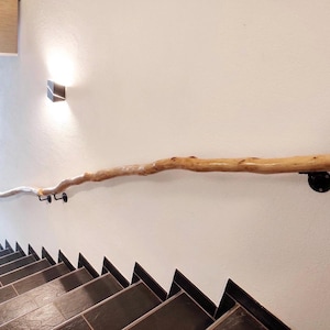 Könnte beinhalten: Ein natürlicher Holzhandlauf an einer weißen Wand. Der Handlauf besteht aus einem einzelnen, hellfarbigen Ast, der von schwarzen Metallhalterungen getragen wird. Die Treppe ist darunter sichtbar.