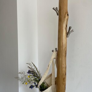 May include: A rustic wooden coat rack with three silver hooks. A canvas tote bag hangs from one hook, filled with wildflowers. The bag has a printed design of a house and text.