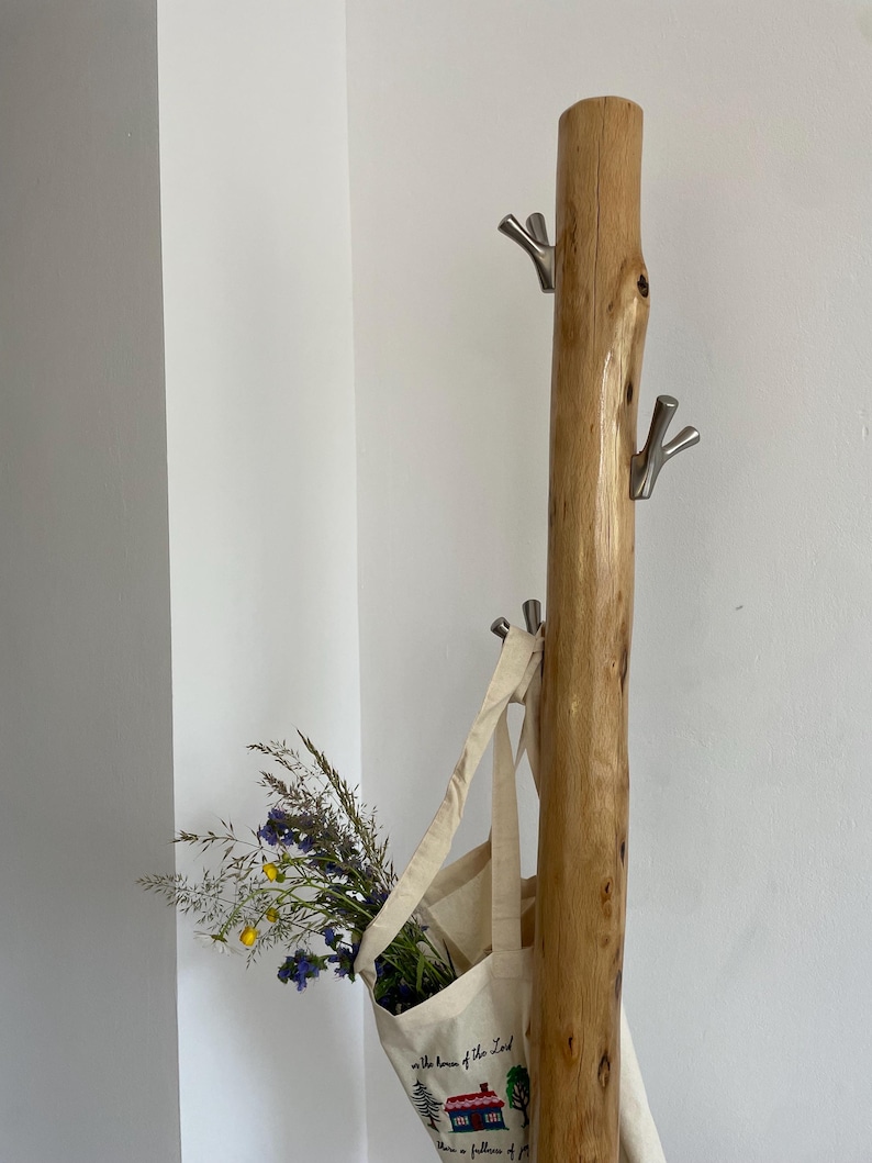 May include: A rustic wooden coat rack with three silver hooks. A canvas tote bag hangs from one hook, filled with wildflowers. The bag has a printed design of a house and text.