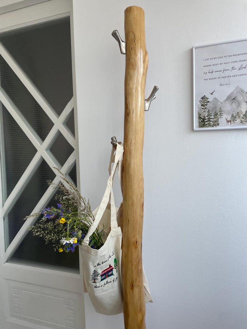 May include: A natural wood coat rack with three silver hooks. A canvas tote bag with a house graphic and text hangs from the bottom hook. The bag is filled with wildflowers. A framed print hangs on the wall.