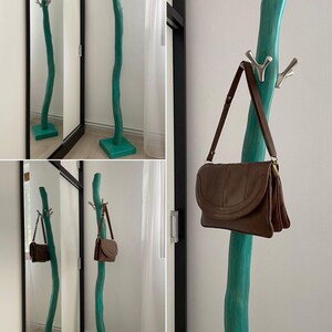 May include: A teal-colored, free-standing coat rack with a natural, curved design. The rack has multiple silver hooks and a square base. A brown leather shoulder bag hangs from one of the hooks.