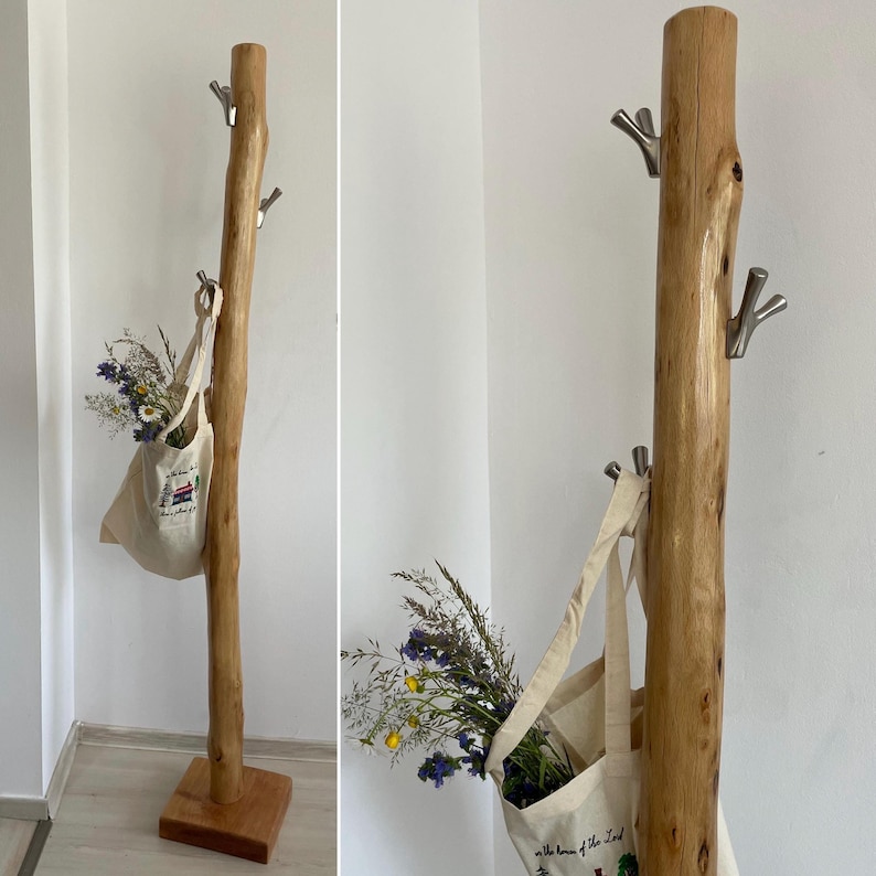 May include: A rustic wooden coat rack with a natural tree trunk design. It features three metal hooks and a square base. A canvas tote bag filled with flowers hangs from one hook, adding a touch of nature. The rack is a light brown color.