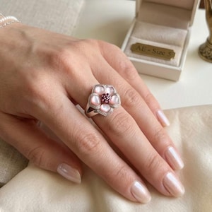 May include: A silver ring featuring a flower design with pink petals, a central cluster of small red stones, and a halo of clear crystals. The ring is on a hand with polished nails.