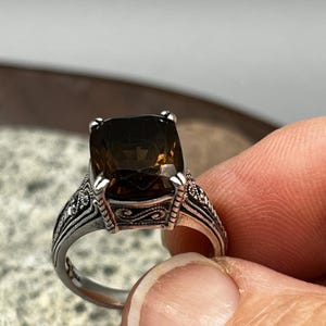 May include: A silver ring with a large, square, smoky brown gemstone. The ring has intricate filigree detailing on the band and around the gemstone setting. The ring is held up by a person's fingers.