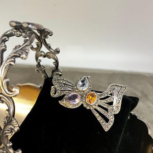 May include: A silver-toned brooch in a bow shape, adorned with marcasite stones and three colorful gemstones: light blue, purple, and orange. The brooch is displayed on a black velvet surface.