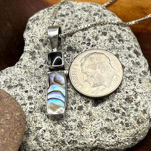 May include: A silver pendant necklace with a rectangular abalone shell and black stone pendant. The abalone shell has iridescent colors of blue, green, and purple. The pendant is next to a US dime.
