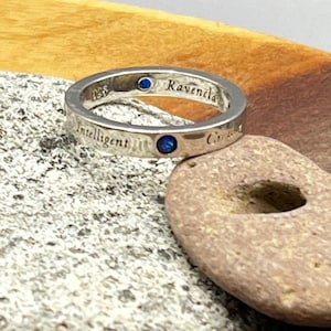 May include: A silver ring with the words "Intelligent" and "Ravenclaw" engraved on it. The ring has two blue gemstones and is set on a gray and brown stone surface.