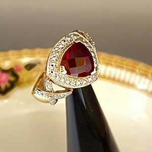 May include: A gold ring with a large, triangular, faceted red gemstone as the centerpiece. The gemstone is surrounded by a halo of smaller, clear stones. The ring's band has a decorative design with additional small stones.