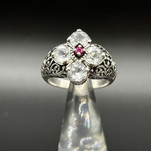 May include: A silver-toned ring featuring a floral design with five clear, round gemstones. A small, red gemstone is set in the center. The ring has an ornate, filigree band.