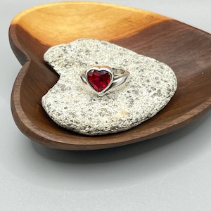 May include: A silver ring featuring a faceted, heart-shaped red gemstone set in a smooth metal band. The ring is positioned on a textured, light gray stone, which rests inside a polished, dark brown wooden bowl shaped like a heart.