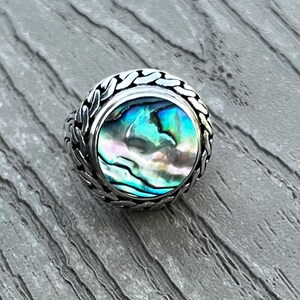 May include: A silver ring featuring a round, iridescent abalone shell inlay. The metal band is crafted with a textured, braided-style pattern surrounding the central stone, which displays swirling shades of blue, green, and pink mother-of-pearl.