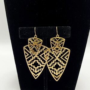 May include: A pair of gold-colored earrings with a geometric design. The earrings have a layered, openwork design with a triangular shape. The intricate patterns include zigzags, triangles, and other shapes. The earrings are displayed on a black stand.