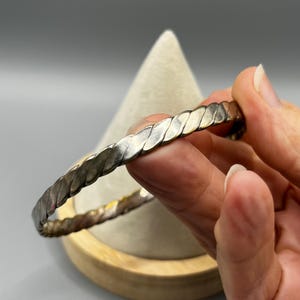 May include: Silver-toned bangle bracelet with a braided design. The bracelet is held in a hand, showcasing its texture and shine. The bracelet's design is made of interlocking, slightly curved metal pieces.