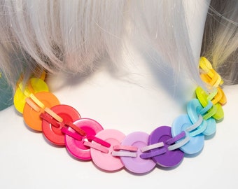 Può includere: Una collana di bottoni arcobaleno con bottoni gialli, arancioni, rossi, rosa, blu chiaro, blu scuro e viola. La collana è su un busto di manichino bianco.